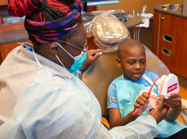 A picture of a dentist using a model of a mouth to show a young boy how to brush his teeth.